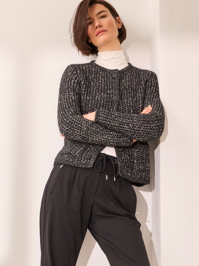 Eine Frau trägt eine grob gestrickte, schwarz-weiß melierte Strickjacke mit großen Knöpfen über einem weißen Rollkragenpullover, kombiniert mit einer schwarzen Hose im modernen, entspannten Stil.