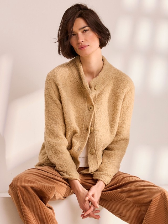 Eine Frau trägt eine beigefarbene, flauschige Strickjacke mit großen Knöpfen über einem hellen Oberteil, kombiniert mit einer braunen Cordhose im warmen, gemütlichen Stil.