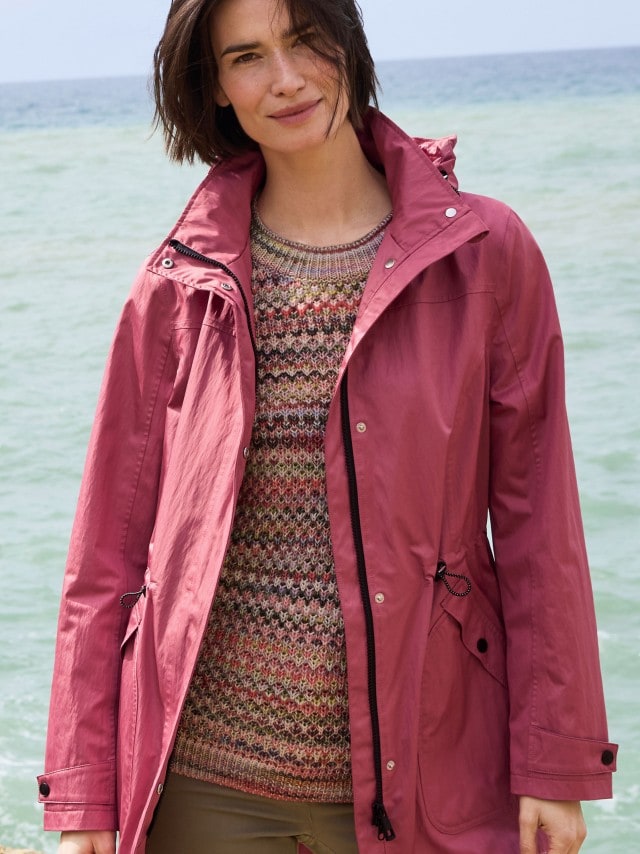 Ein Frau steht am Strand und trägt einen Parka in der Farbe Rosenquarz, darunter einen gemusterten Strickpullover und dazu eine Hose in Khaki.