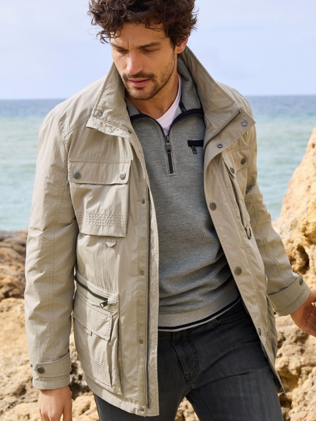 Ein Mann steht an einer Felsküste und trägt die Aquastop Traveller Jacke, darunter einen Troyer und dazu eine Jeans in grau.