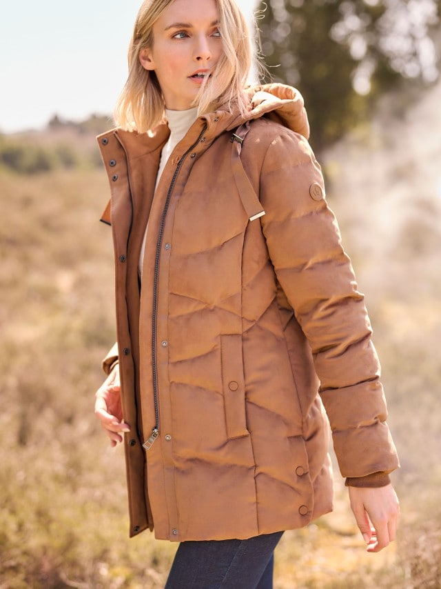 Eine Frau trägt eine mittelbraune, gesteppte Jacke mit Kapuze, Reißverschluss und Druckknöpfen, kombiniert mit einem beigen Rollkragenpullover im modernen, sportlich-eleganten Stil.