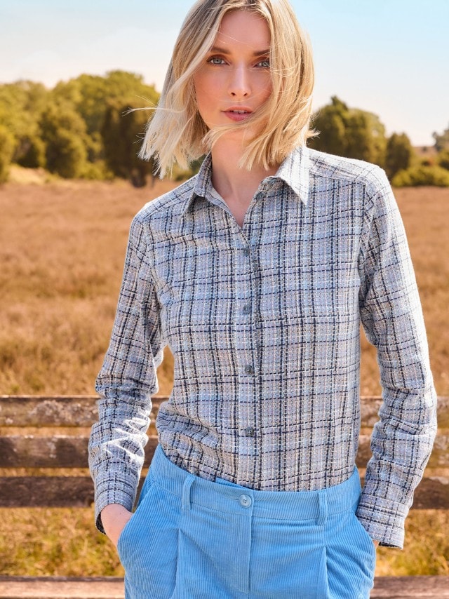 Eine Frau trägt ein langärmliges Hemd mit feinem Karomuster in Grau- und Blautönen, kombiniert mit einer hellblauen Cordhose im stilvollen, modernen Look.