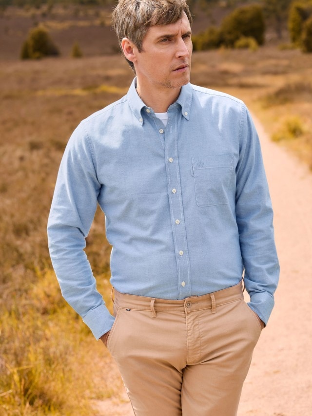Ein Mann geht auf einem Feldweg und trägt ein hellblaues Button-down-Hemd sowie eine beigefarbene Chinohose.