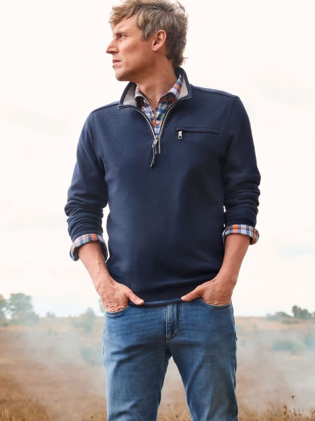 Ein Mann steht im Freien und trägt ein dunkelblaues Langarm-Sweatshirt mit Reißverschluss über einem karierten Hemd sowie eine Jeans.