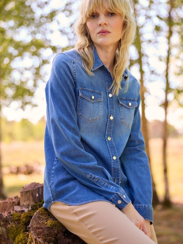 Eine Frau trägt ein klassisches Jeanshemd mit Druckknöpfen und Brusttaschen, kombiniert mit einer beigefarbenen Hose im entspannten, zeitlosen Stil.