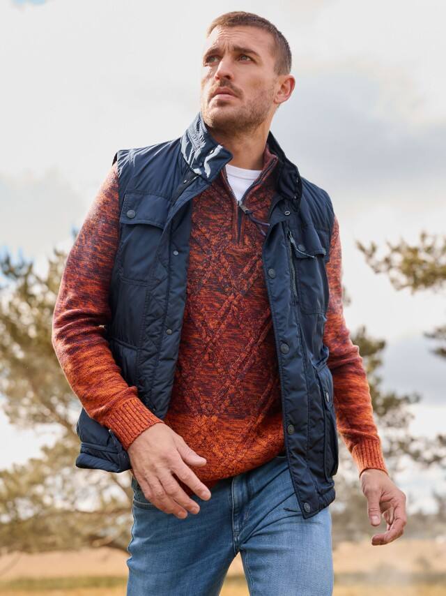 Ein Mann trägt einen orange-roten Strickpullover mit Muster, eine dunkle Steppweste und Jeans – lässiger, herbstlicher Outdoor-Look.