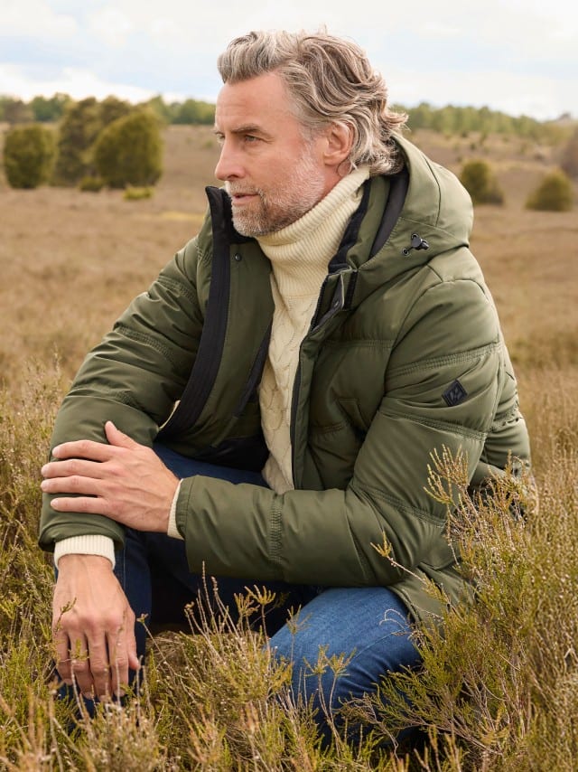 Ein Mann trägt eine olivgrüne Steppjacke über einem cremefarbenen Zopfpullover mit Rollkragen und Jeans; der Look wirkt gemütlich und winterlich.