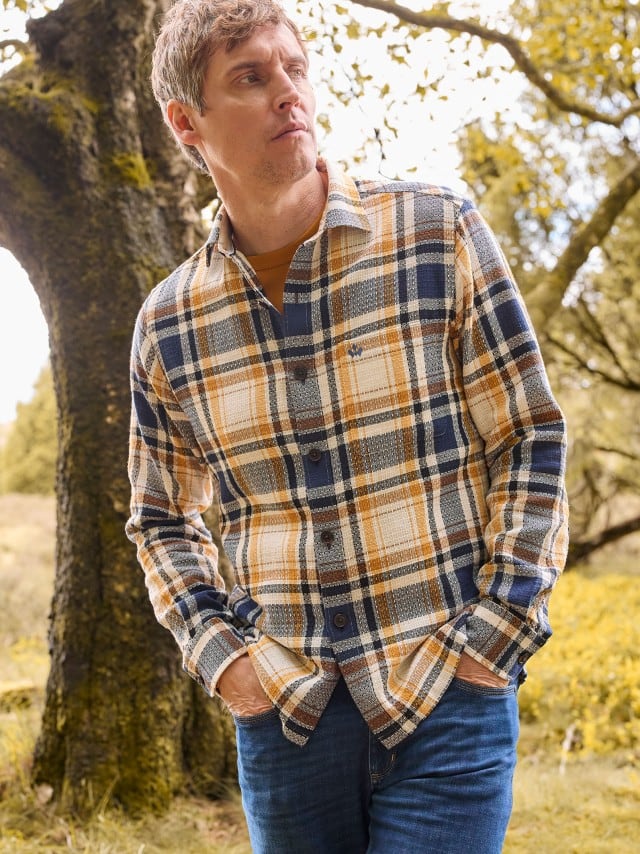 Ein Mann trägt ein kariertes Flanellhemd in Blau-, Weiß- und Ockertönen zu Jeans; der Look wirkt lässig und herbstlich.