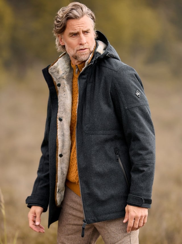 Ein Mann trägt eine dunkelgraue Winterjacke mit heller Innenfütterung über einem senfgelben Strickpullover und karierte Stoffhosen; der Look wirkt warm und stilvoll herbstlich.