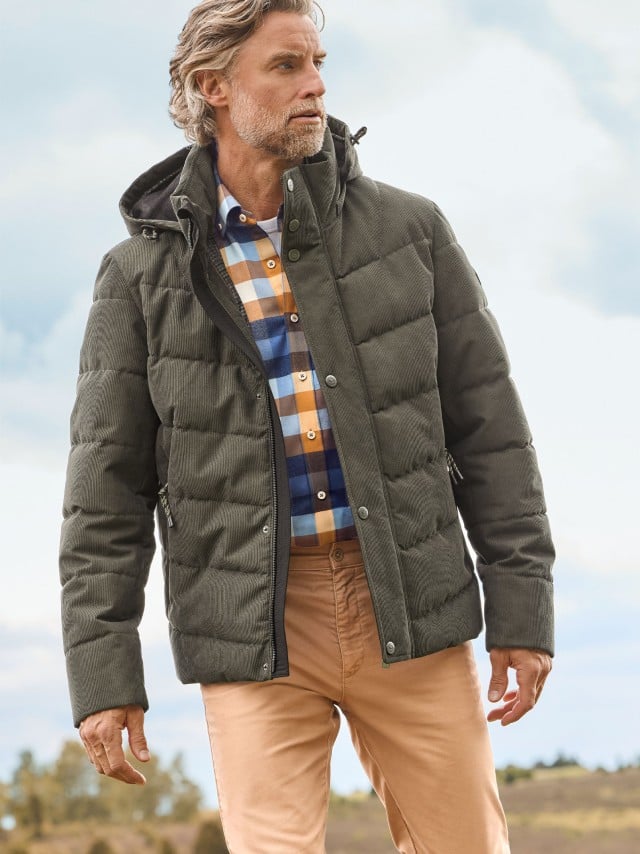 Ein Mann trägt eine olivgrüne Steppjacke über einem karierten Hemd in Blau-, Weiß- und Ockertönen sowie beigefarbene Chinos; der Look wirkt modern und herbstlich.