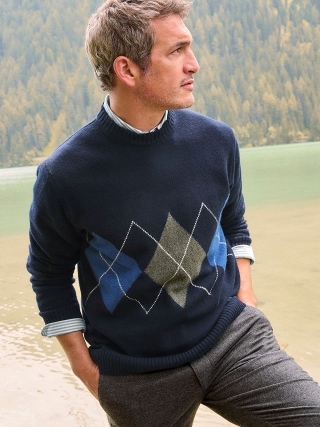 Ein Mann steht vor einem See und trägt den Argyle Pullover in Marine, darunter ein hellblaues Hemd und dazu eine Sport-Flanell Hose in Anthrazit.