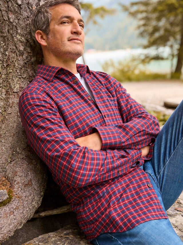 Ein Mann sitzt an einem Baum gelehnt und trägt ein Sturmtief Flanell-Hemd in Karo Bordeaux, darunter ein weißes Shirt und dazu eine Jeans.