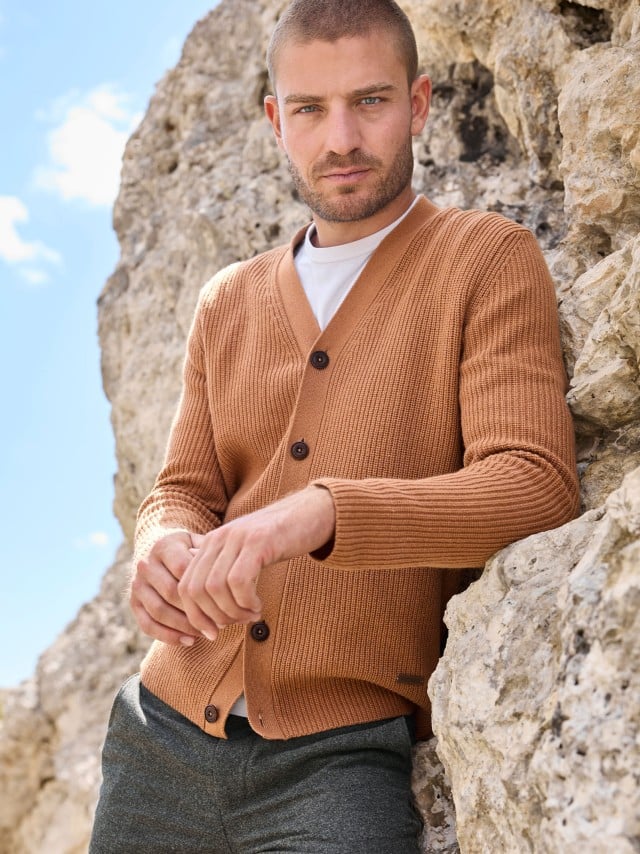 Ein Mann steht an einer Felswand und trägt den Patentstrick-Cardigan in Karamell, darunter ein weißes Shirt und dazu eine dunkele Flanellhose.