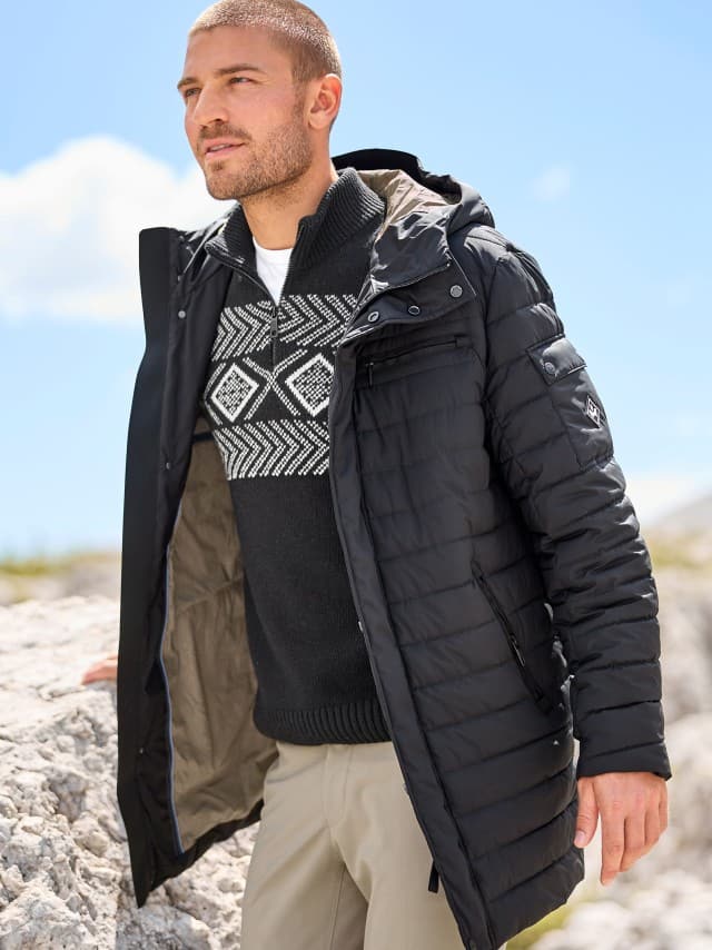 Ein Mann steht vor Felsen und trägt einen Leichtsteppmantel in Schwarz, darunter einen Norweger Troyer und dazu eine helle Hose.