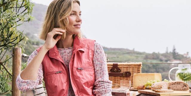 Frau sitzt auf einer Wanderung draußen und trägt eine leichte, farbenfrohe Weste und schaut in die Ferne.