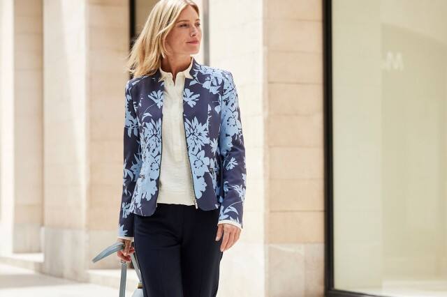 Frau in einem blau gemustertem Blazer und Tasche läuft an einer Fensterfront entlang.