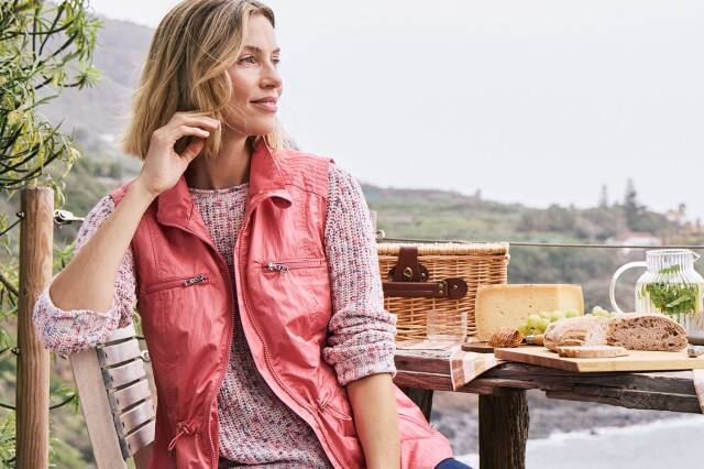 Frau sitzt auf einer Wanderung draußen, trägt eine leichte, farbenfrohe Weste und schaut in die Ferne.