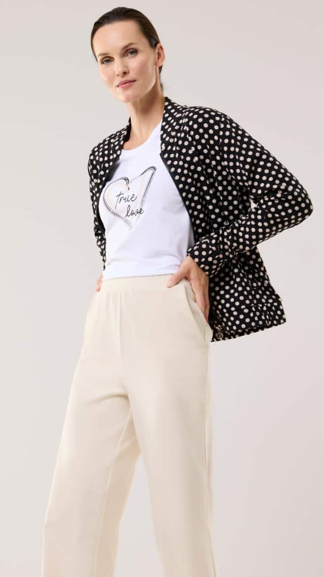 Eine Frau trägt eine schwarz‑weiß gepunktete Jacke über einem weißen T‑Shirt mit Herzmotiv und heller, weiter Hose. Der Look wirkt modern, lässig und leicht elegant.