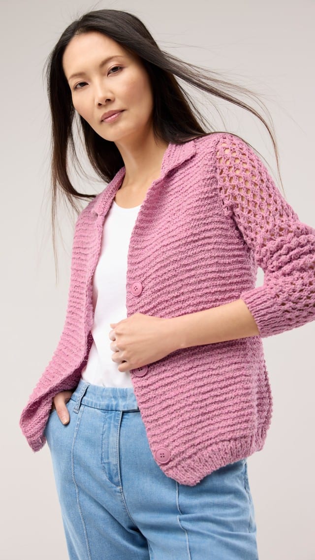 Eine Frau trägt eine rosafarbene Strickjacke mit durchbrochenen Ärmeln über einem weißen Oberteil und einer hellblauen Hose. Eine Frau trägt eine rosafarbene Strickjacke mit durchbrochenen Ärmeln über einem weißen Oberteil und einer hellblauen Hose.