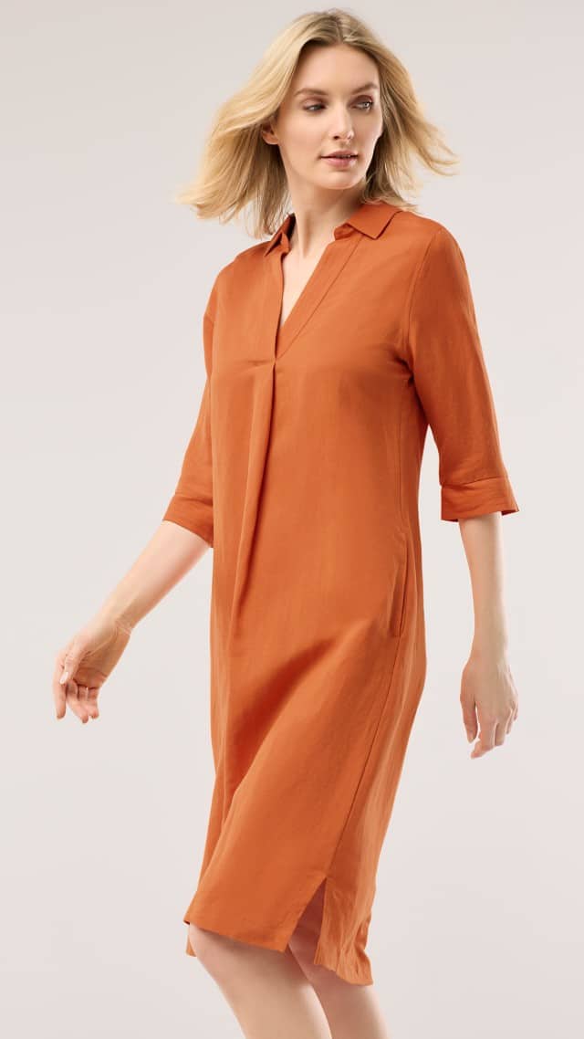 Eine Frau trägt ein knielanges, orangefarbenes Kleid mit halblangen Ärmeln und läuft leicht seitwärts. Eine Frau trägt ein knielanges, orangefarbenes Kleid mit halblangen Ärmeln und läuft leicht seitwärts.