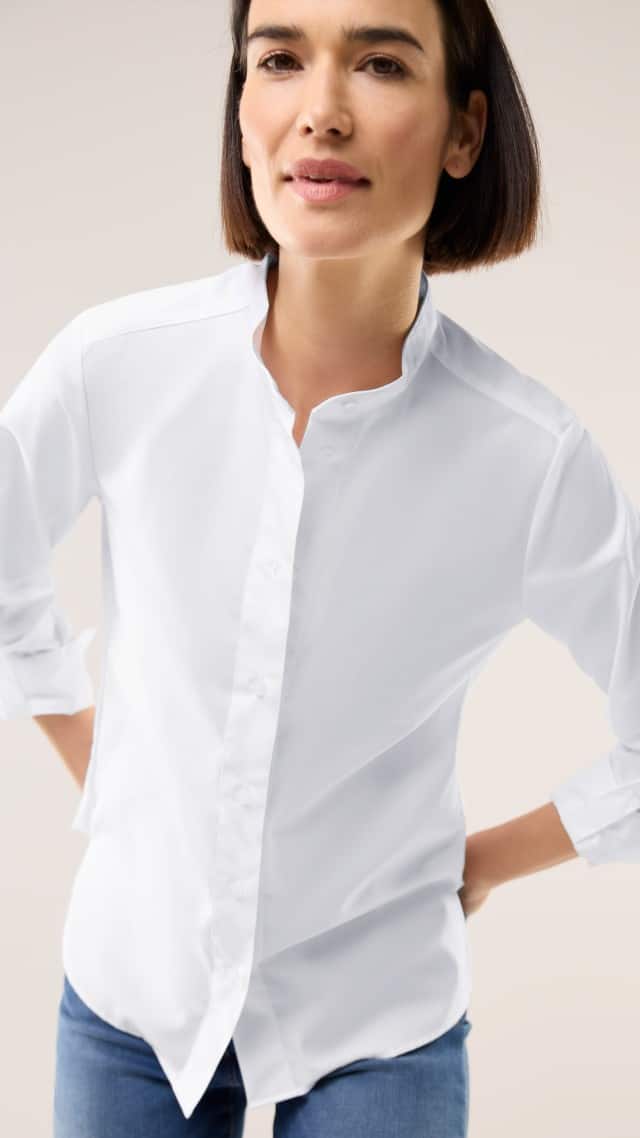 Eine Frau trägt ein schlichtes, weißes Hemd mit Stehkragen und aufgerollten Ärmeln. Der Stil wirkt klassisch, elegant und zeitlos.