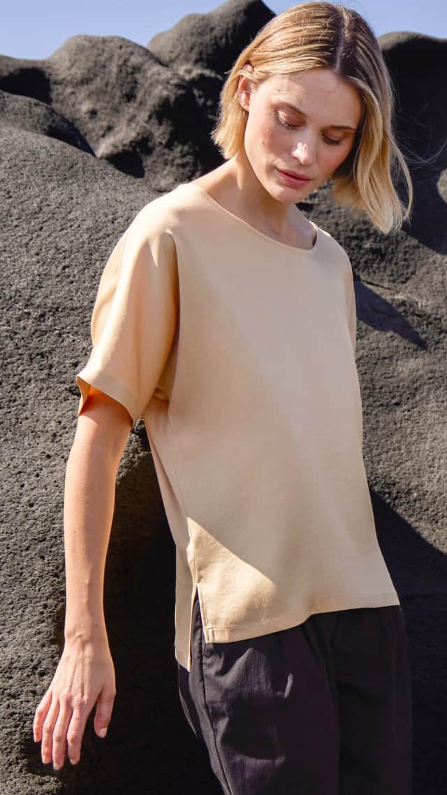 Eine Frau steht vor einem Felsen und trägt ein Blusenshirt aus Satin in der Farbe Sand, dazu eine schwarze Hose.