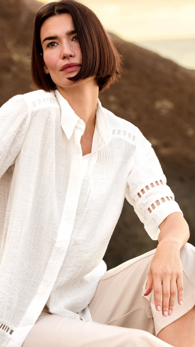 Eine Frau sitzt an einem Felsen am Strand und trägt eine weiße Statement Bluse, dazu eine Hose in Beige.