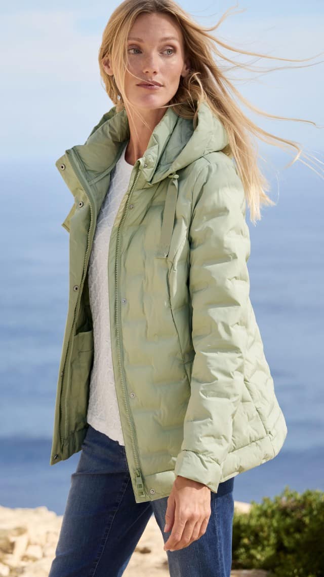 Eine Frau steht im Freien und trägt eine mintgrüne Steppjacke zu einem weißen Oberteil und Jeans. Eine Frau steht im Freien und trägt eine mintgrüne Steppjacke zu einem weißen Oberteil und Jeans.