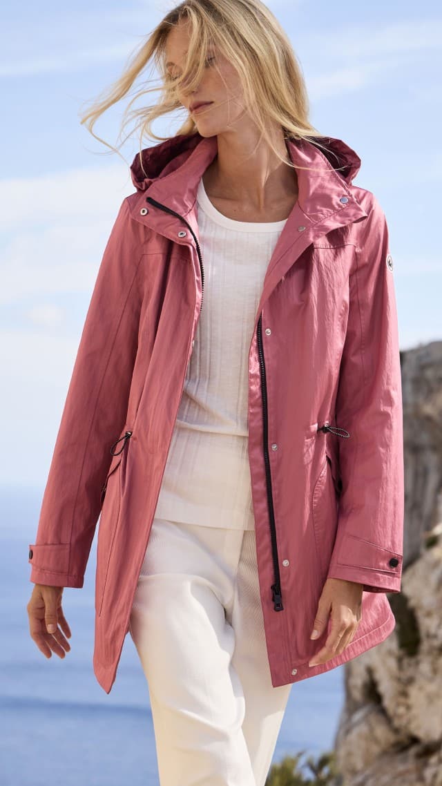 Eine Frau steht im Freien und trägt eine rosafarbene Jacke über einem weißen Oberteil und einer weißen Hose. Eine Frau steht im Freien und trägt eine rosafarbene Jacke über einem weißen Oberteil und einer weißen Hose.