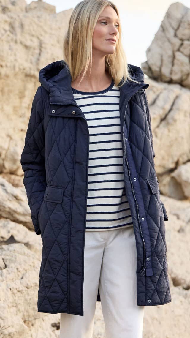 Eine Frau steht vor Felsen und trägt einen dunkel gesteppten Mantel über einem gestreiften Oberteil. Eine Frau steht vor Felsen und trägt einen dunkel gesteppten Mantel über einem gestreiften Oberteil.