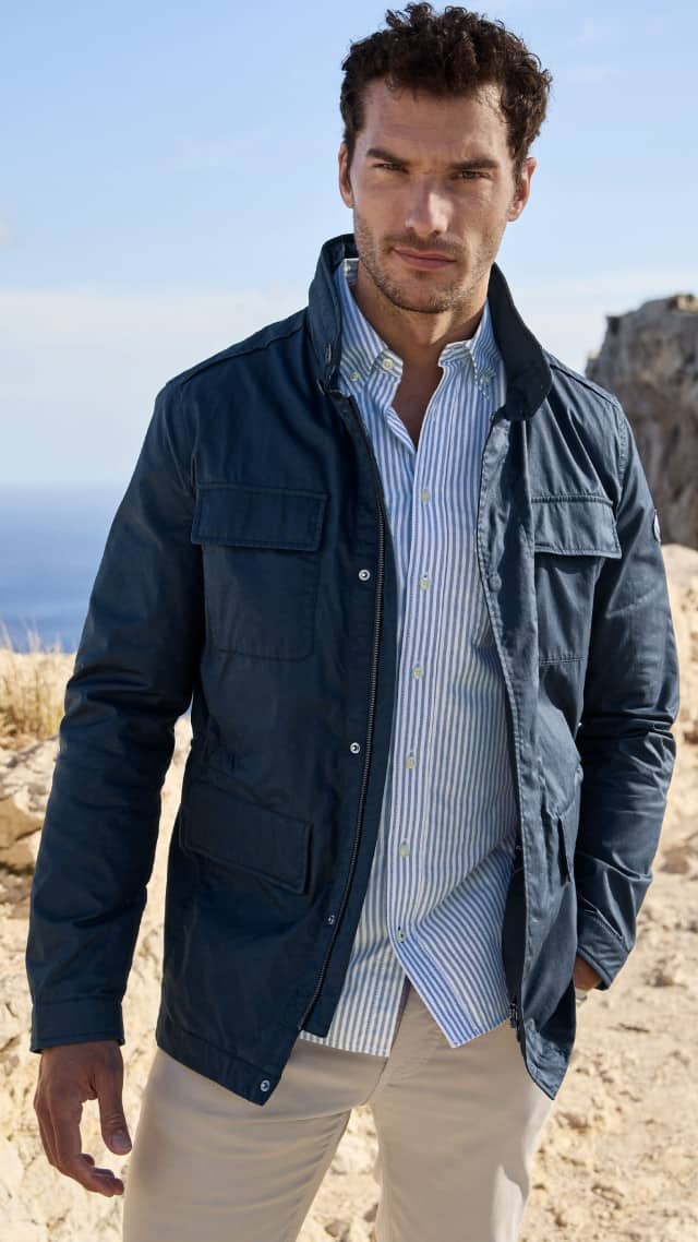 Ein Mann steht in einer hellen Felsenlandschaft und trägt eine dunkelblaue Jacke über einem gestreiften Hemd.