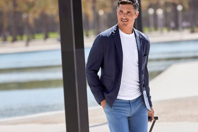 Mann in einem Business Casual Outfit und einem Koffer steht in einer Parkanlage.