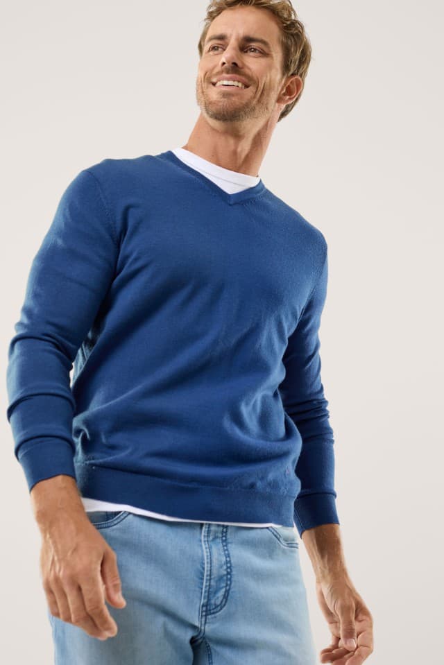 Ein Mann trägt einen blauen Pullover mit V-Ausschnitt, darunter ein weißes Shirt und dazu eine hellblaue Jeans.