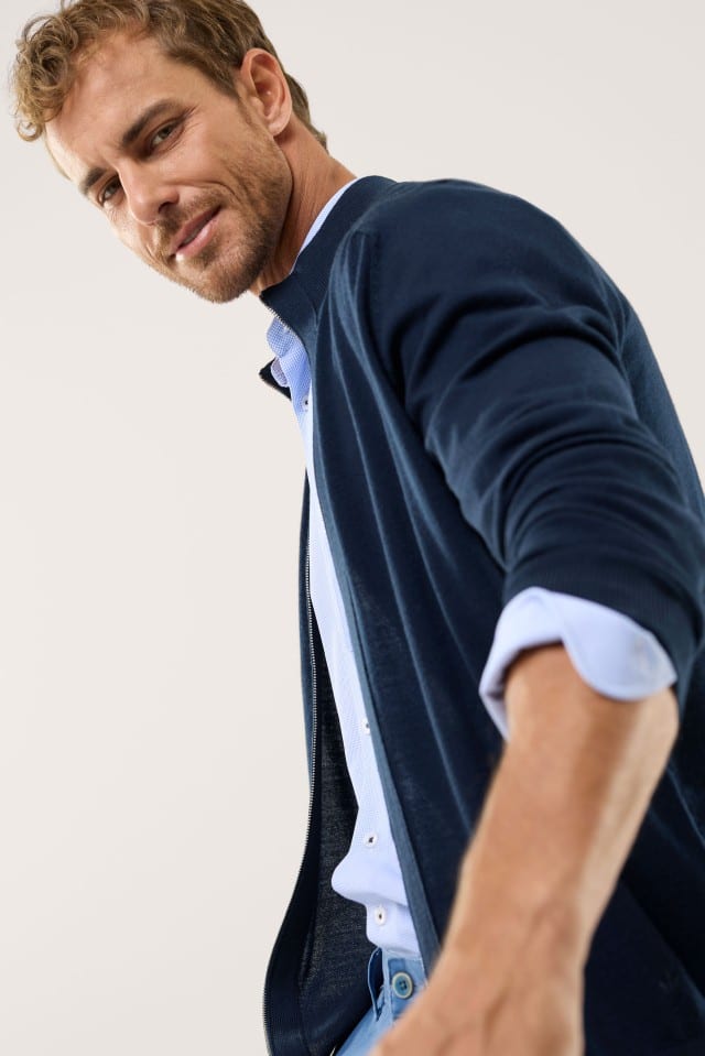 Ein Mann trägt eine Zip-Strickjacke in Marine, darunter ein hellblaues Hemd und dazu eine Jeans.