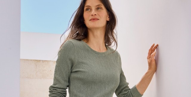 Eine Frau steht an einer weißen Wand und trägt ein langärmliges, strukturiertes Oberteil in einem dezenten Grünton.
