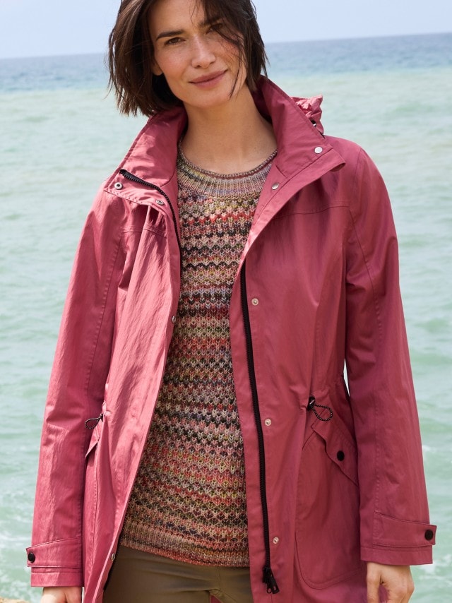Ein Frau steh am Strand und trägt den Aquastop Parka in der Farbe Rosenquarz, darunter einen gemusterten Strickpullover.