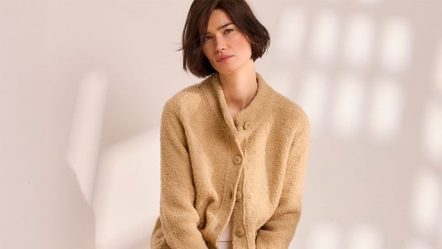 Eine Frau vor einem neutralen Hintergrund trägt die Baby Alpaka Strickjacke in der Farbe Sand.