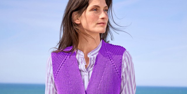 Eine Frau trägt eine gestrickte, lila Weste über einer lila-weiß gestreiften Bluse. Der Stil wirkt modern und farbenfroh.