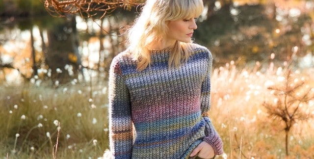 Ein blonde Frau steht auf einer herbstlichen Wiese, schaut zur Seite und trägt einen Grobstrick-Pullover mit Farbverlauf.