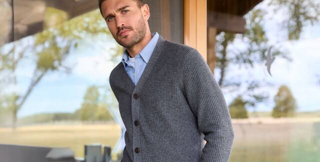 Ein Mann steht vor einer großen Fensterfront und trägt ein hellblaues Hemde, darüber einen dunklen Strick-Cardigan. Ein Mann steht vor einer großen Fensterfront und trägt ein hellblaues Hemde, darüber einen dunklen Strick-Cardigan.