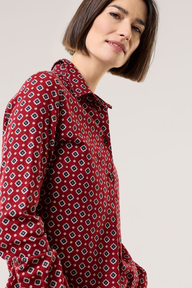 Eine Frau trägt ein langärmliges rotes Hemd mit kleinem, geometrischem Muster in Blau, Weiß und Schwarz, das einen eleganten und modernen Stil hat.