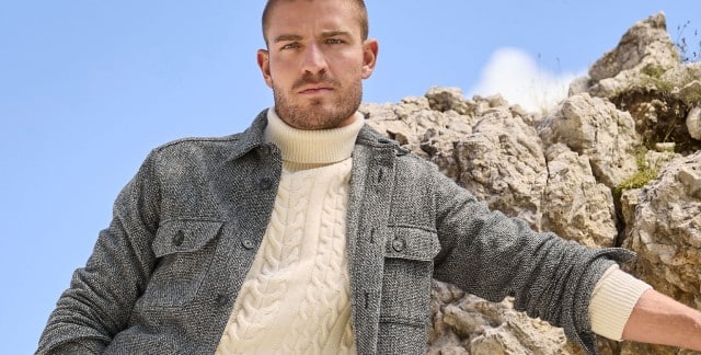 Ein Mann steht vor Felsen und trägt eine Hemd-Jacke in Schwarz/Weiß, darunter einen Rollkragen-Pullover mit Zopfmuster in Wollweiß. Ein Mann steht vor Felsen und trägt eine Hemd-Jacke in Schwarz/Weiß, darunter einen Rollkragen-Pullover mit Zopfmuster in Wollweiß.