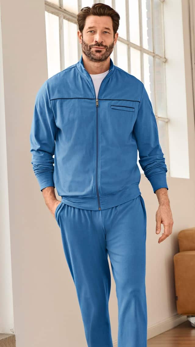 Ein Mann trägt einen hellblauen Freizeitanzug mit Zip‑Jacke und passender Hose über einem weißen T‑Shirt. Der Look wirkt sportlich, bequem und lässig.
