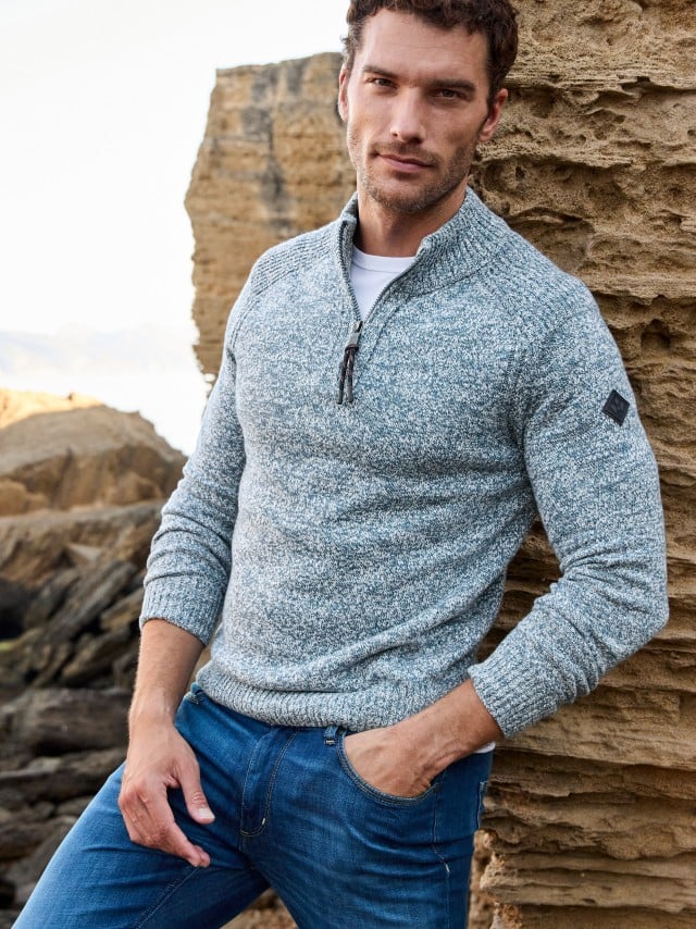 Ein Mann lehnt an einer Felswand und trägt einen hellgrau melierten Troyer-Pullover sowie eine Jeans.
