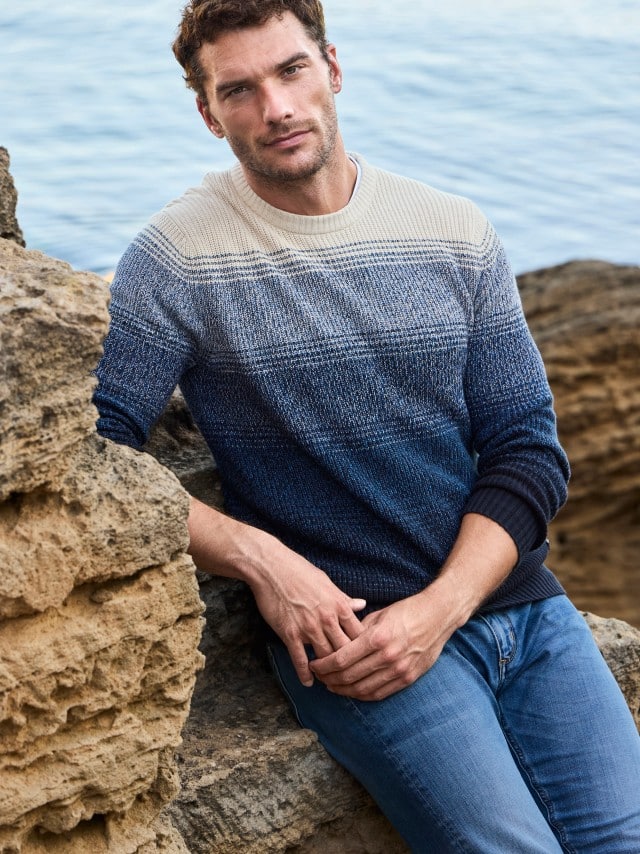 Ein Mann sitzt zwischen Felsen am Wasser und trägt einen blau-beigen Strickpullover sowie eine Jeans.