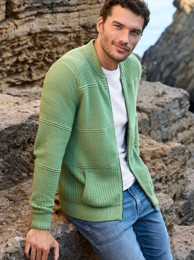 Ein Mann sitzt auf Felsen und trägt eine hellgrüne Strickjacke über einem weißen T‑Shirt sowie eine Jeans.