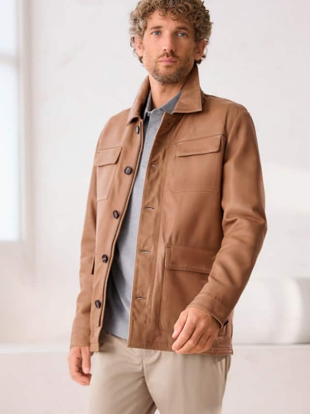 Ein Mann trägt die Multitaschen Lederjacke in der Farbe Cognac, darunter ein graues Polo-Shirt und dazu eine Hose in Beige. 