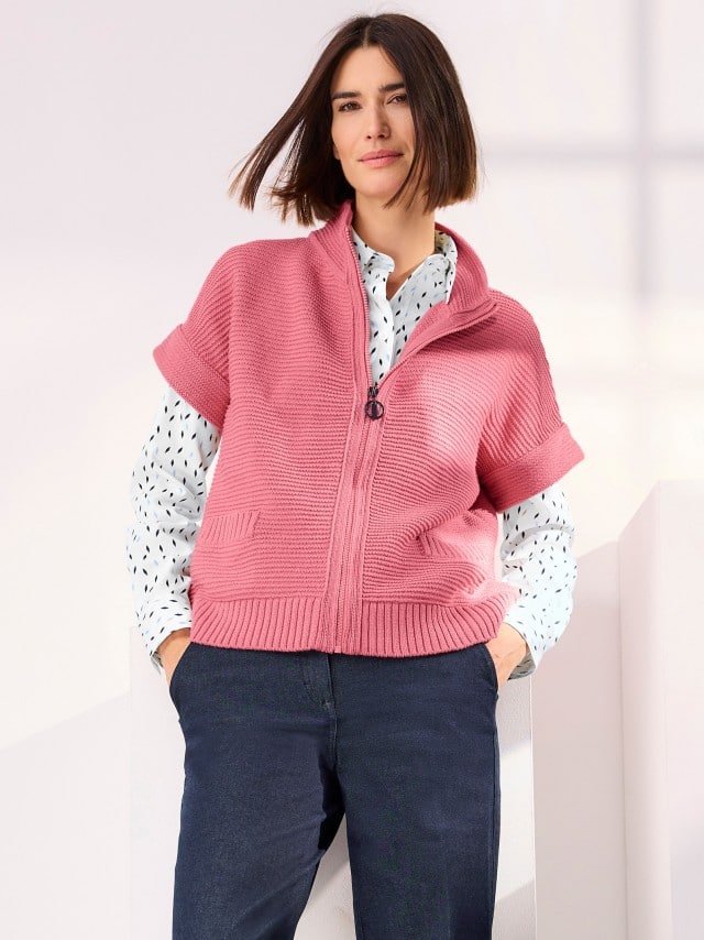 Frau trägt eine rosafarbene Strickjacke mit Reißverschluss über einer weißen Bluse mit schwarzem Muster und dunkler Hose – legerer, moderner Freizeit-Look.