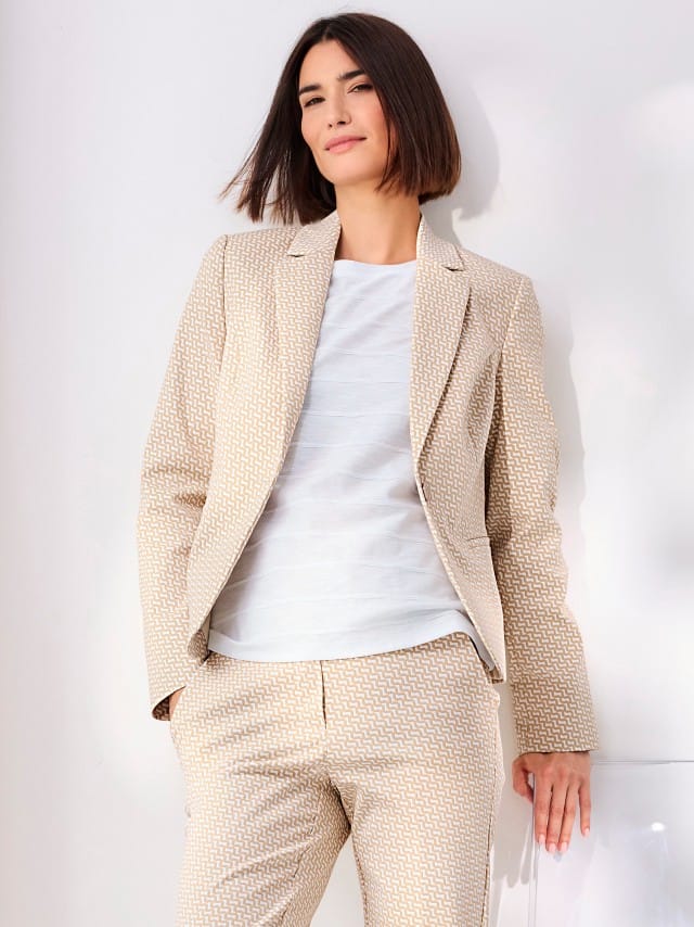 Frau trägt einen beigefarbenen Hosenanzug mit feinem Muster über einem weißen Shirt – moderner, stilvoller Business-Look mit leichter Note.