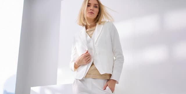 Eine blonde Frau trägt einen weißen Seersucker Blazer, mit passender Weste und Hose, dazu ein Baumwollshirt mit Streifenstruktur in Beige. 